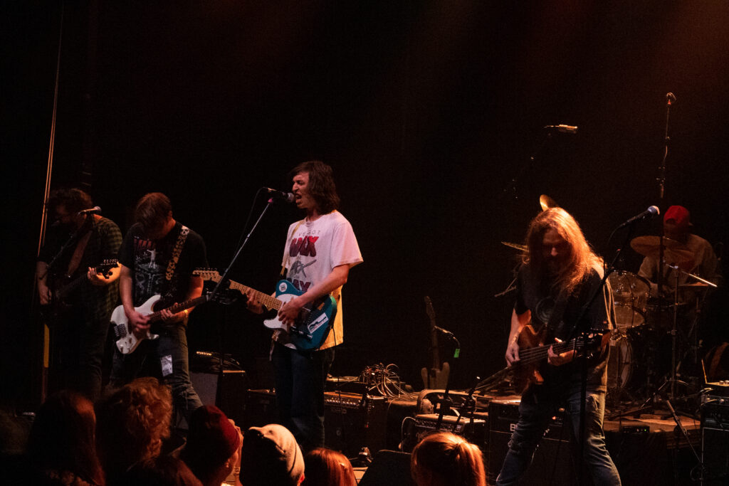 A band playing a song, with the lead singer in a white t-shirt playing guitar and seemingly singing very loudly into a microphone